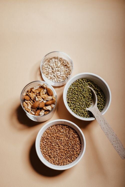 A top-down view of four small bowls containing mix-ins: oats, almonds with some nuts, green lentils or mung beans, and crushed brown grains, with a wooden spoon in one bowl. How to mix protein powder without clump.